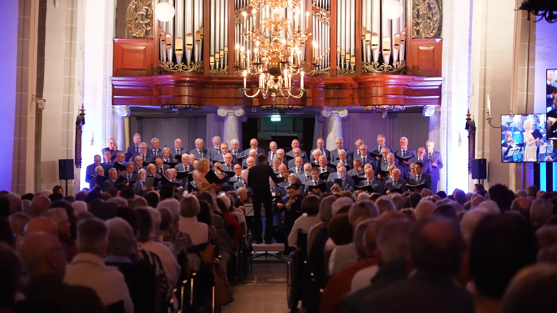 Mannenkoor, orkest en publiek in een kerk, met het orgel op de achtergrond